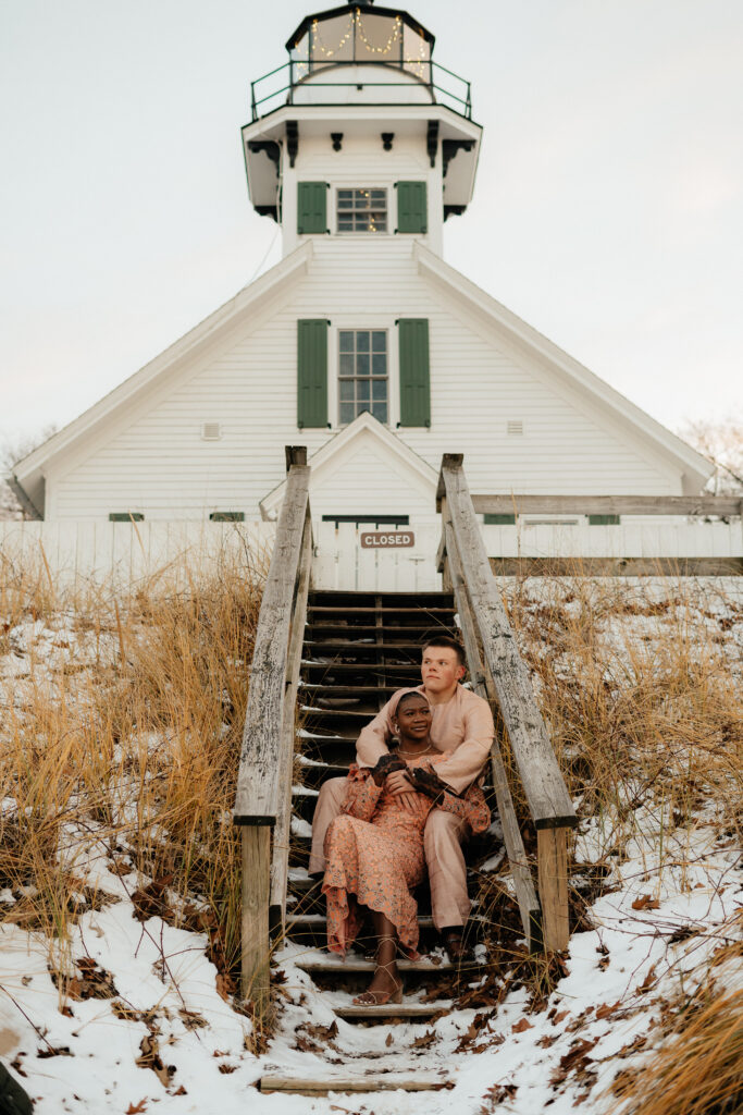 A couple sits on the stairs of a popular Traverse City Lighthouse for their engagement photos 