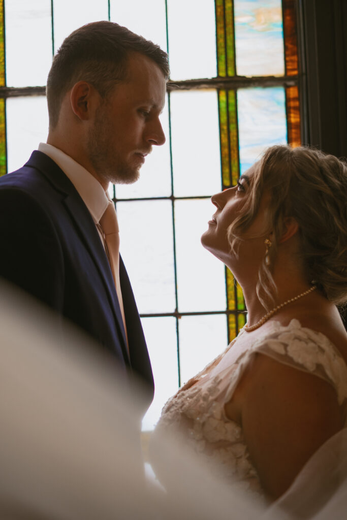 A couple gazes into each others eyes in front of a stained glass window at The Chapel at New Richmond in Saugatuck, Michigan