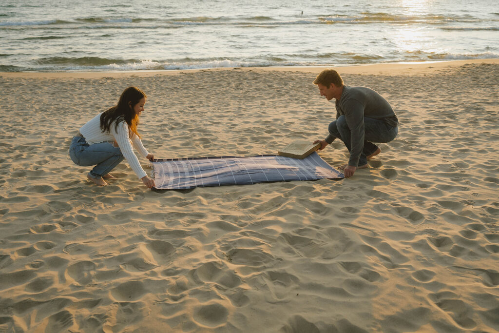 couple stretch out a blanket to start their beach day photoshoot