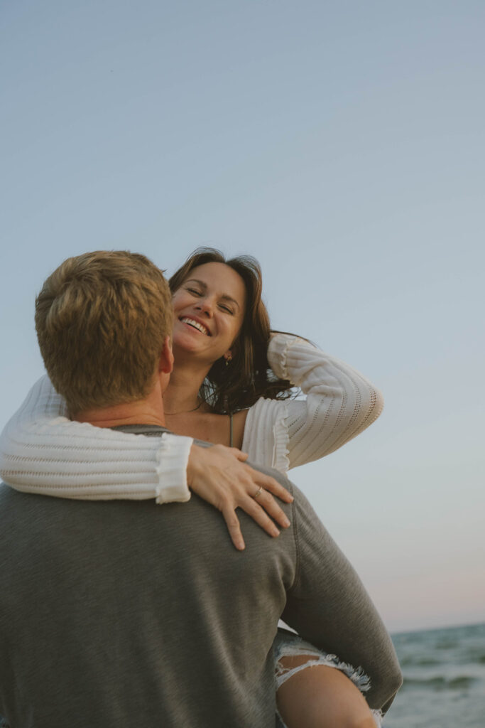 future bride smiles at the camera as her man holds her on the Lake Michigan shore