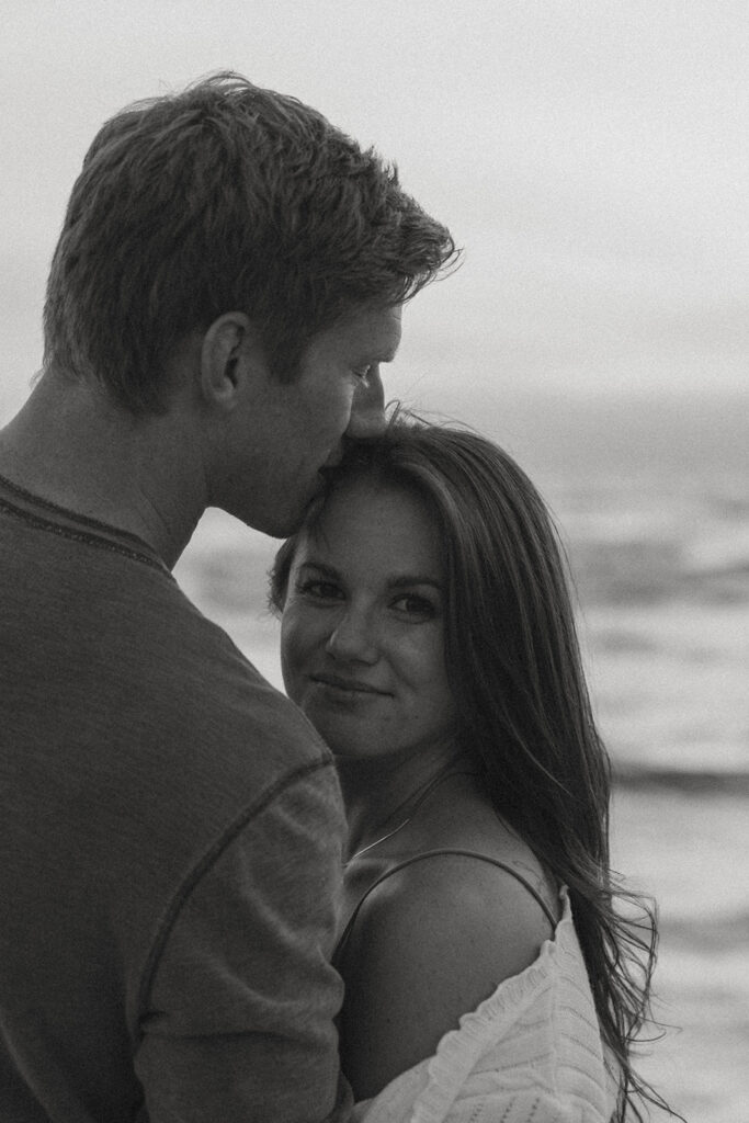future bride smiles at the camera as her man holds her on the Lake Michigan shore