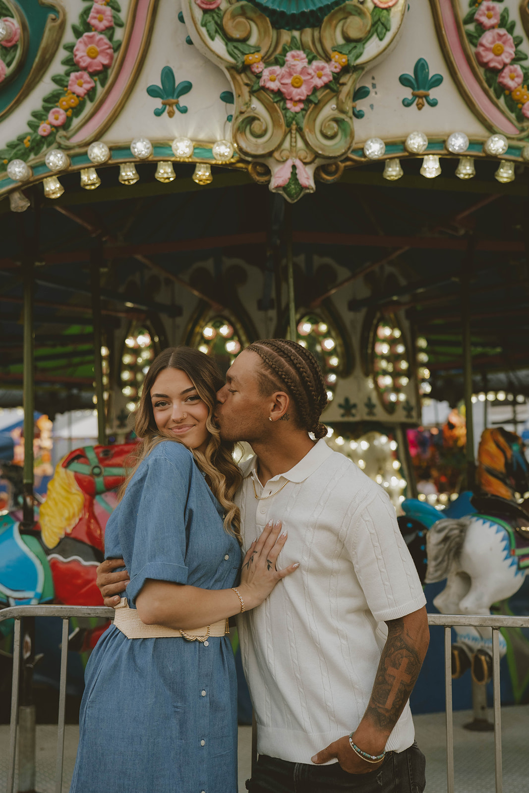 couple share a romantic kiss on the cheek in front of the merry go round during their state fair photoshoot