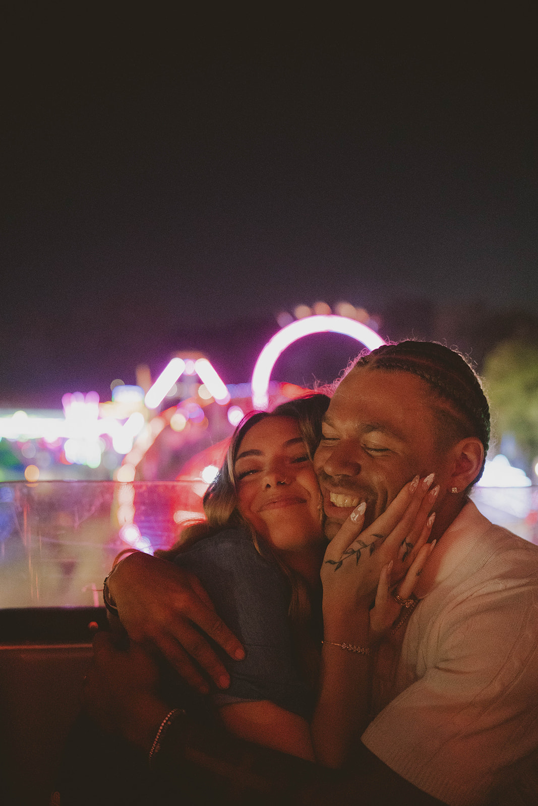 couple embrace and smile as the fair lights them up in a warm pink light