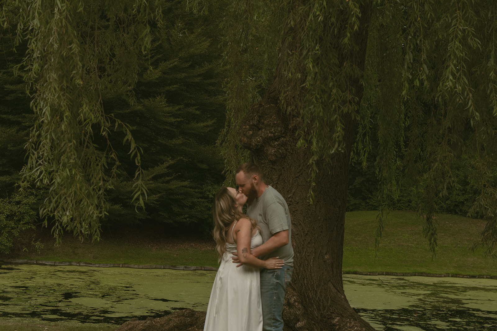 couple share a kiss in front of the tree standing over the pond 